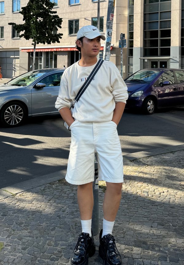 Vit långärmad skjorta med logotyp, vita shorts, svarta chunky sneakers och en vit keps. Står bredvid en gatulampa, urban bakgrund.