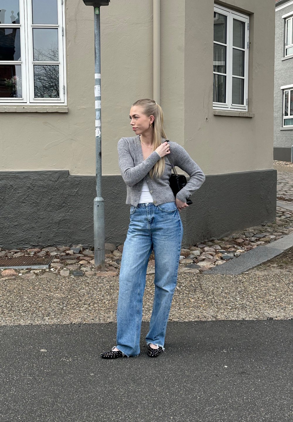 Cardigan grigio lavorato a maglia, top bianco, jeans a gamba larga blu chiaro e scarpe slip-on nere con perline. Sullo sfondo di un edificio di colore neutro.