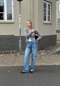 Cardigan grigio lavorato a maglia, top bianco, jeans a gamba larga blu chiaro e scarpe slip-on nere con perline. Sullo sfondo di un edificio di colore neutro.