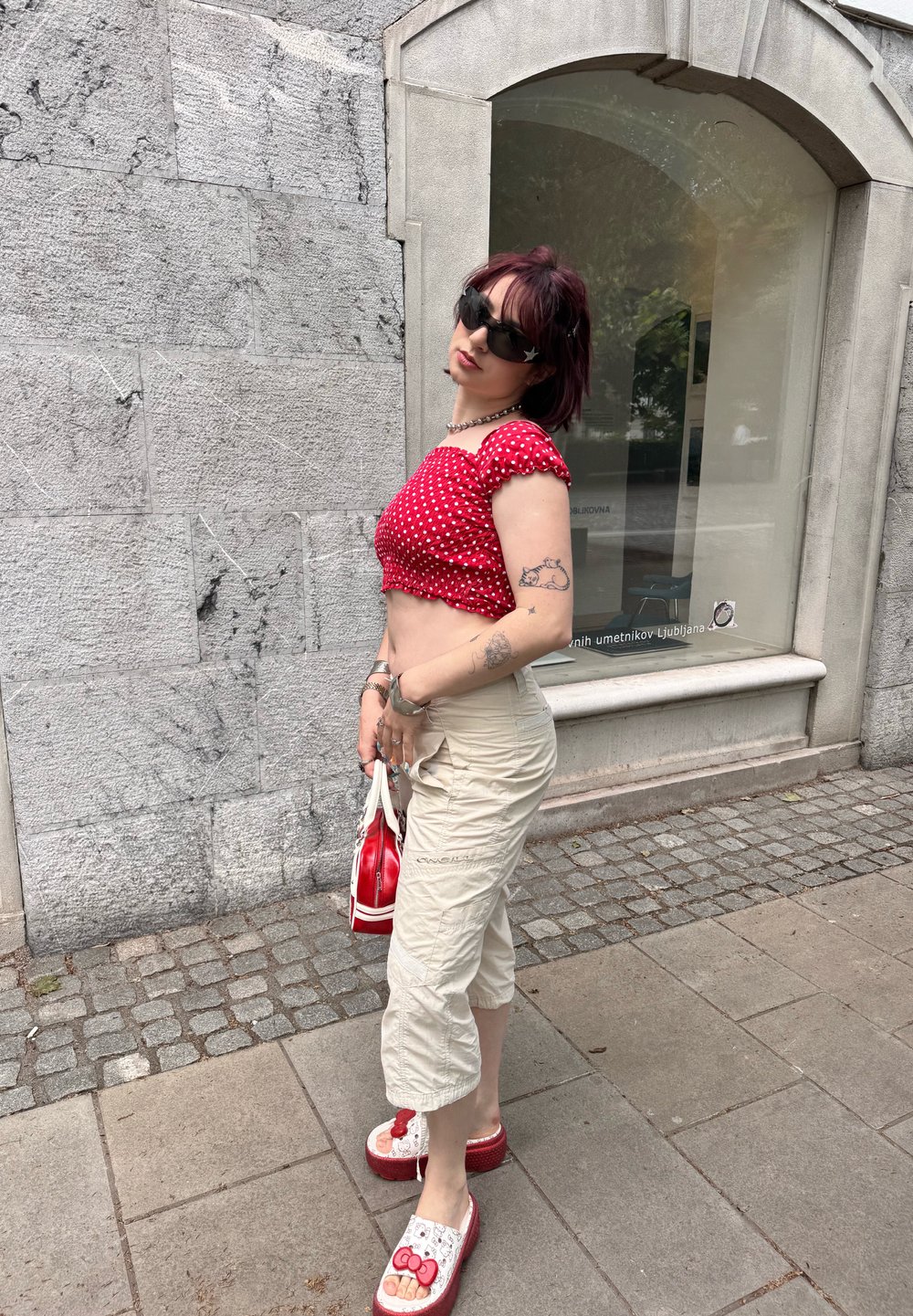 Top crop vermelho com bolinhas, calças cargo bege, tamancos vermelhos e brancos com um design da Hello Kitty, segurando uma pequena bolsa vermelha.