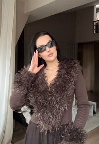 Pulover maro cu dungi, cu guler pufos din blană artificială și accente pe mâneci; asortat cu ochelari de soare închiși la culoare; interior, fundal neutru.