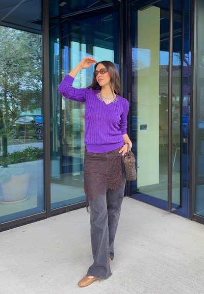 Mujer con suéter morado, falda con encaje sobre jeans, gafas de sol y bolso de diseñador, parada junto a puertas de vidrio al aire libre.