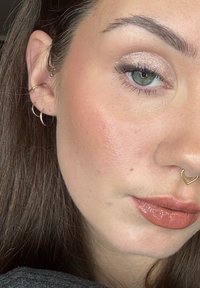 Lähikuva vaaleanruskeahiuksisesta kasvoista, joissa on kultaiset renkaanmuotoiset korvakorut, sydämenmuotoinen nenänreikäkoru ja kiiltävä luonnollisen sävyinen huulipuna.