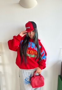Sweat à capuche rouge surdimensionné avec texte graphique bleu et vert, pantalon de survêtement gris avec impression bleue, casquette rouge et noire, et petit sac à main rouge.