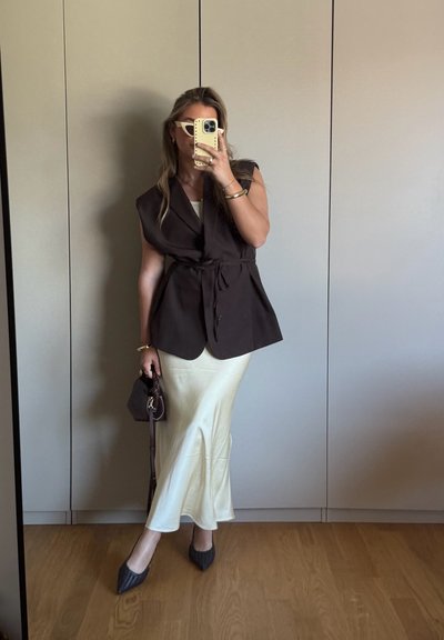 Chaleco sin mangas marrón con cinturón, falda midi de satén crema, tacones de punta oscura y texturizada, sosteniendo un pequeño bolso oscuro, de pie contra un fondo neutro.