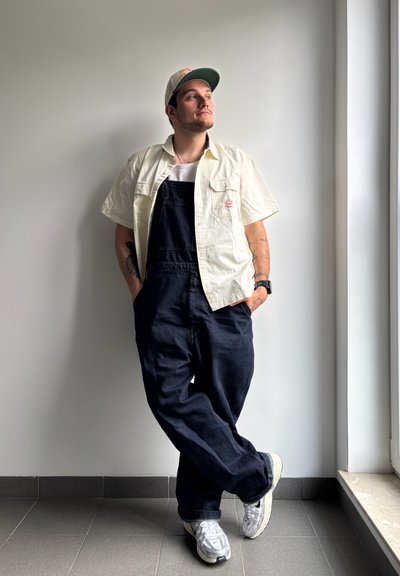 Camisa blanca de manga corta, overoles azul oscuro, zapatillas grises. Persona de pie contra una pared, con los brazos cruzados y una gorra. Fondo neutro.