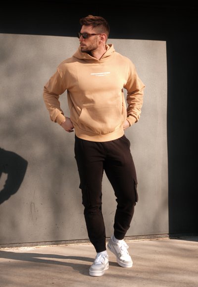 Sudadera beige con capucha y bolsillo frontal, pantalones cargo negros y zapatillas blancas. Los detalles notables incluyen texto impreso en la sudadera.