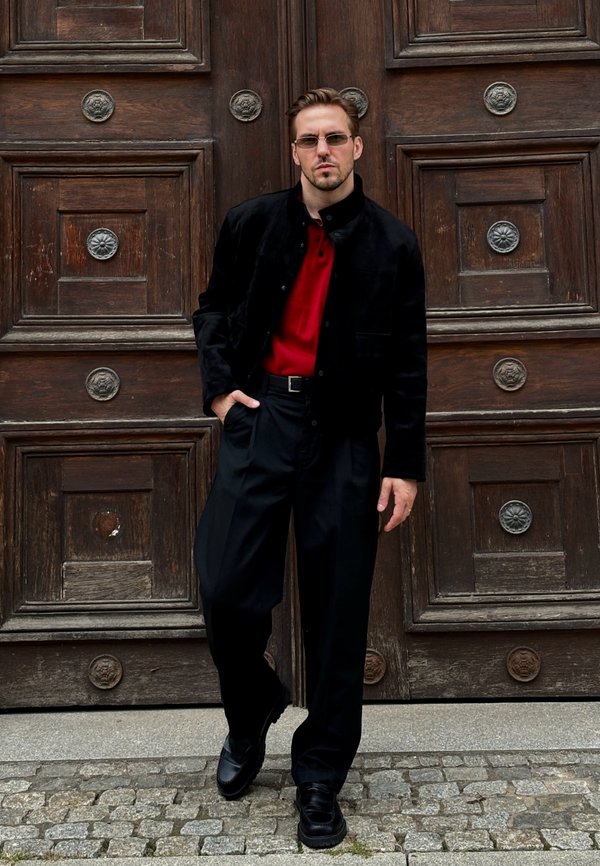 Hombre con chaqueta negra, camisa roja, pantalones negros y gafas de sol, de pie con una mano en el bolsillo frente a una gran puerta de madera con detalles decorativos de metal.