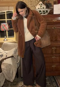 Manteau en fausse fourrure marron, pull crème et pantalon large sombre. Sac à main marron, textures subtiles et matériaux superposés. Intérieur avec éclairage chaud.
