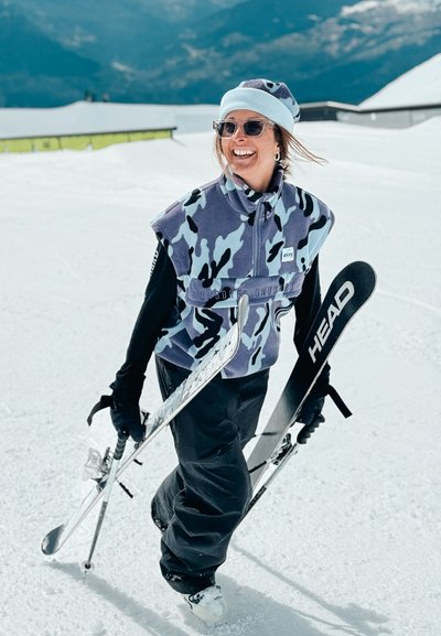 Mujer sonriente con polar azul de camuflaje, pantalones negros y sombrero camina sobre la nieve sosteniendo esquís y bastones, con montañas de fondo.