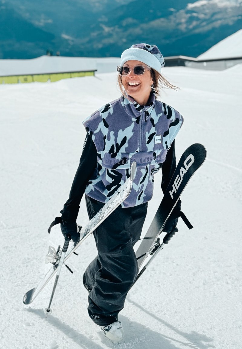 Lachende vrouw in blauwe camouflagefleece, zwarte broek en hoed loopt op sneeuw terwijl ze ski's en stokken vasthoudt met bergen op de achtergrond.