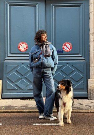 Ein Modell trägt eine blaue Jeansjacke und Jeans, einen gestreiften Schal, und steht neben einem Hund auf einer Straße mit blauen Türen und roten Schildern im Hintergrund.