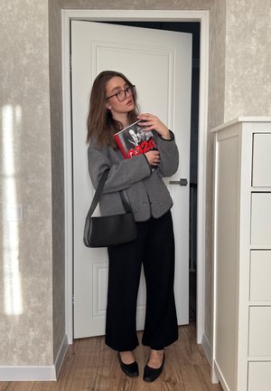 Mujer con gafas, chaqueta gris, pantalones y zapatos negros, sosteniendo un libro con cubierta roja y negra y un bolso negro en el interior.