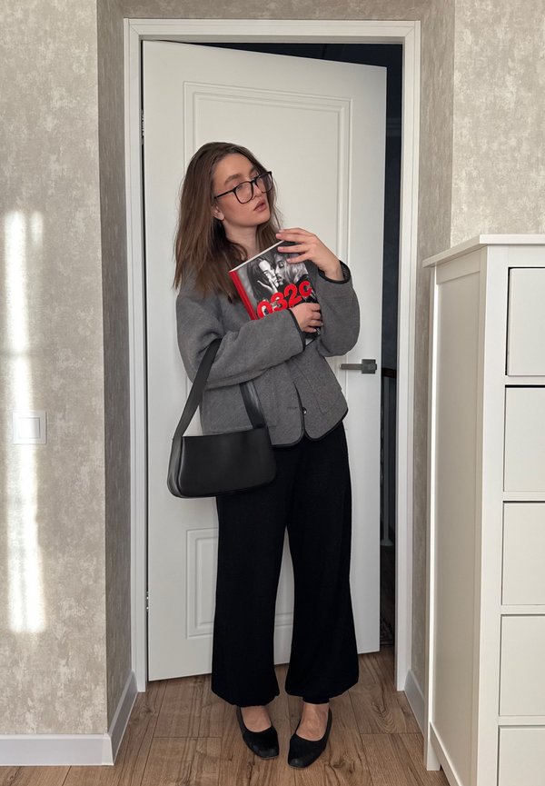 Vrouw met bril, grijze jas, zwarte broek en schoenen, die binnen een boek met een rood-zwarte omslag en een zwarte tas vasthoudt.