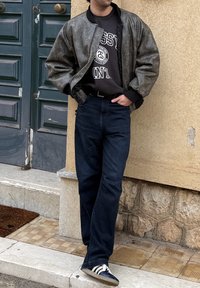 Άντρας που ακουμπά σε υφή μπεζ τοίχο, φορώντας γκρι μπουφάν bomber, μαύρο φούτερ με σχέδιο, σκούρο τζιν και μαύρα-λευκά αθλητικά παπούτσια.