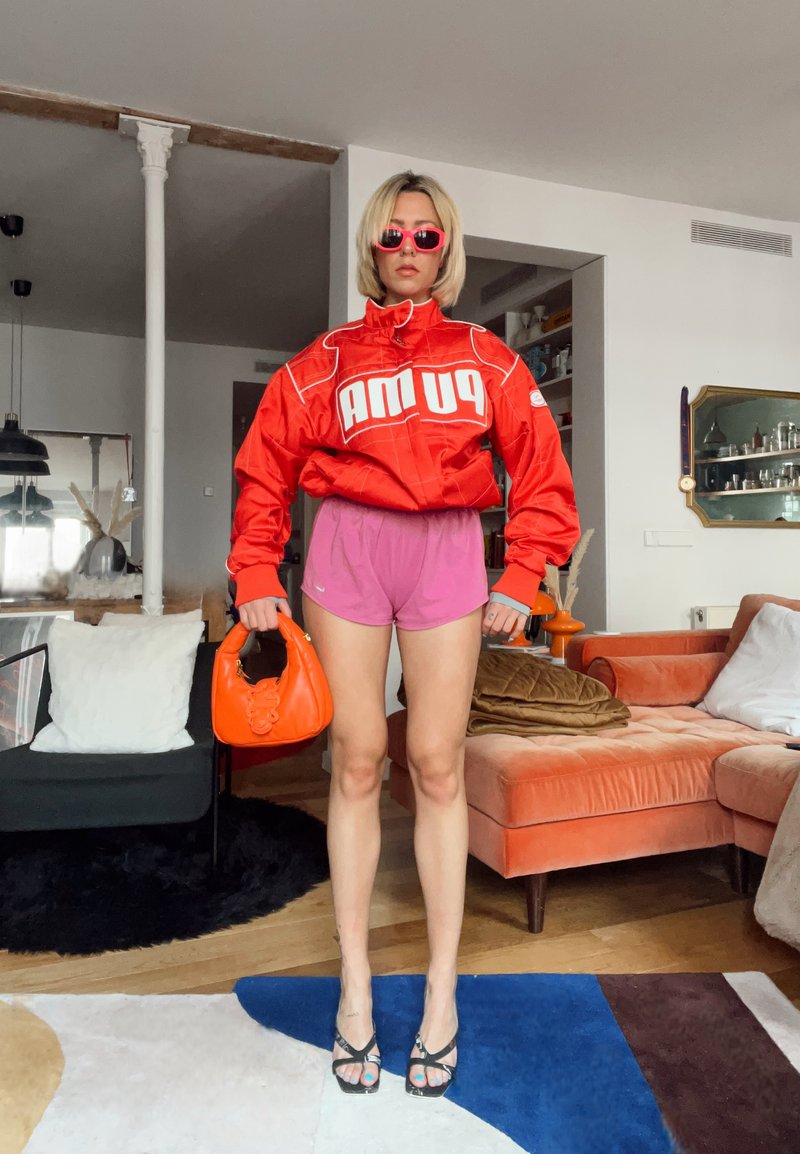Femme aux cheveux blonds coupés au carré, portant une veste rouge Puma, un short rose, des lunettes de soleil rouges, des talons noirs à lanières, tenant un sac à main orange dans un salon.