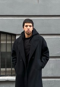 Jeune homme aux cheveux courts et foncés portant un long manteau noir et une chemise noire, debout contre un mur de béton gris avec une fenêtre à barreaux.