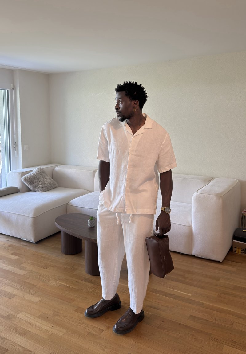 Homme en tenue en lin blanche tenant un sac en cuir marron se tient dans un salon moderne avec un canapé blanc et un sol en bois.