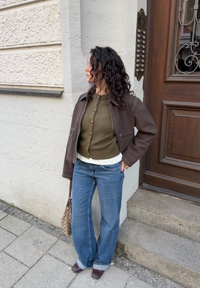 Mujer con cabello rizado, chaqueta marrón y cárdigan verde, jeans azules, sosteniendo un bolso con estampado, de pie junto a la entrada de un edificio en la acera.