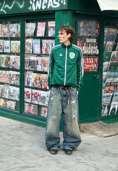 Chaqueta verde de Adidas con rayas blancas, jeans de mezclilla con detalles desgastados, de pie en una acera cerca de un quiosco de revistas.