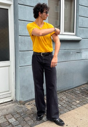 Camiseta amarilla de cuello en V, jeans anchos negros, cinturón negro y zapatos negros. Cabello rizado; gafas de sol puestas, de pie junto a una pared gris y una ventana.