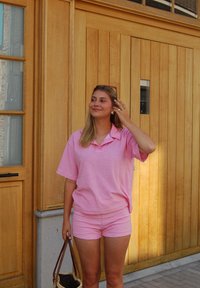 Rosa randig oversized polo tröja och matchande shorts. Mjukt tyg, avslappnad passform. Brun handväska med texturerad yta. Träbakgrund.