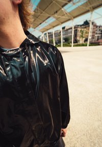 Čierna lesklá bunda s kapucňou, s reflexným povrchom a sťahovanými manžetami. Strih je voľný, zobrazený od ramena nahor.