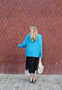 Niebieski, oversize'owy sweter, czarna spódnica z koronkową lamówką, jasna torebka z ćwiekami, na tle czerwonej ściany w romby na brukowanej kostce.