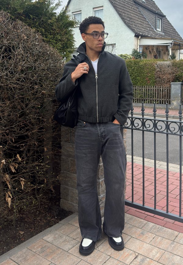 Νέος άνδρας με γυαλιά, φορώντας σκούρο μπουφάν και τζιν, κρατώντας μια μαύρη τσάντα, στέκεται δίπλα σε μεταλλική περίφραξη έξω από ένα σπίτι.