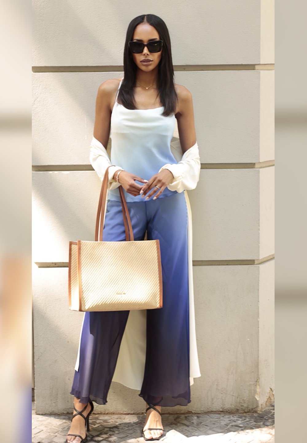 Hellblaues Ombre-Top kombiniert mit fließenden blauen Hosen. Eine weiße Außenschicht, schwarze Sonnenbrille und eine strukturierte beigefarbene Tasche mit braunen Griffen.