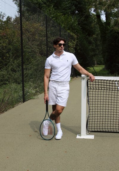 Camiseta polo blanca y pantalones cortos, zapatillas blancas, sosteniendo una raqueta de tenis verde. Cerca de malla y red al fondo, superficie de la cancha de tenis visible.