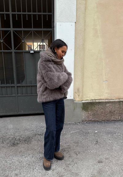 Chaqueta de pelo sintético en gris claro, corte oversize, con mangas anchas y cuello alto. Combinada con jeans de color azul oscuro y calzado marrón.