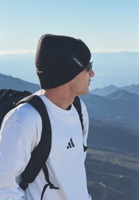 Homme portant un bonnet noir, des lunettes de soleil, un sweat-shirt Adidas blanc et un sac à dos noir regarde un paysage montagneux sous un ciel dégagé.