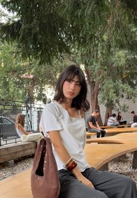 Haut blanc à manches courtes avec un décolleté carré, associé à un pantalon large gris. Sac marron avec de grands trous et des accents métalliques. Cadre naturel.