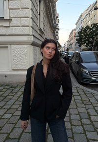 Młoda kobieta w czarnym żakiecie i dżinsach stoi na brukowanej ulicy, gdzie parkują samochody, w otoczeniu zabytkowych budynków o zmierzchu.