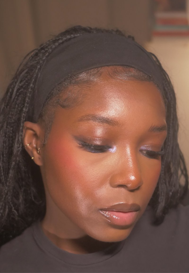 Primapplikation einer Person mit dunklem geflochtenem Haar, die ein schwarzes Stirnband trägt. Merkmale sind die betonte Haut, ein subtiler Rouge-Ton und ein dunkler, geschwungener Eyeliner.