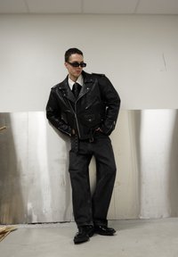 Veste de motard en cuir noir avec des fermetures éclair argentées, portée sur une chemise blanche et une cravate. Un jean en denim foncé et des chaussures noires complètent la tenue.