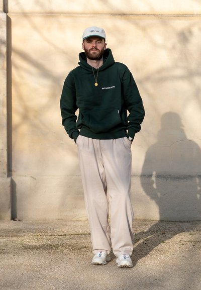 Sudadera verde oscuro con bolsillo frontal y texto blanco, pantalones de pliegues beige, y zapatillas blancas con detalles, de pie contra una pared.