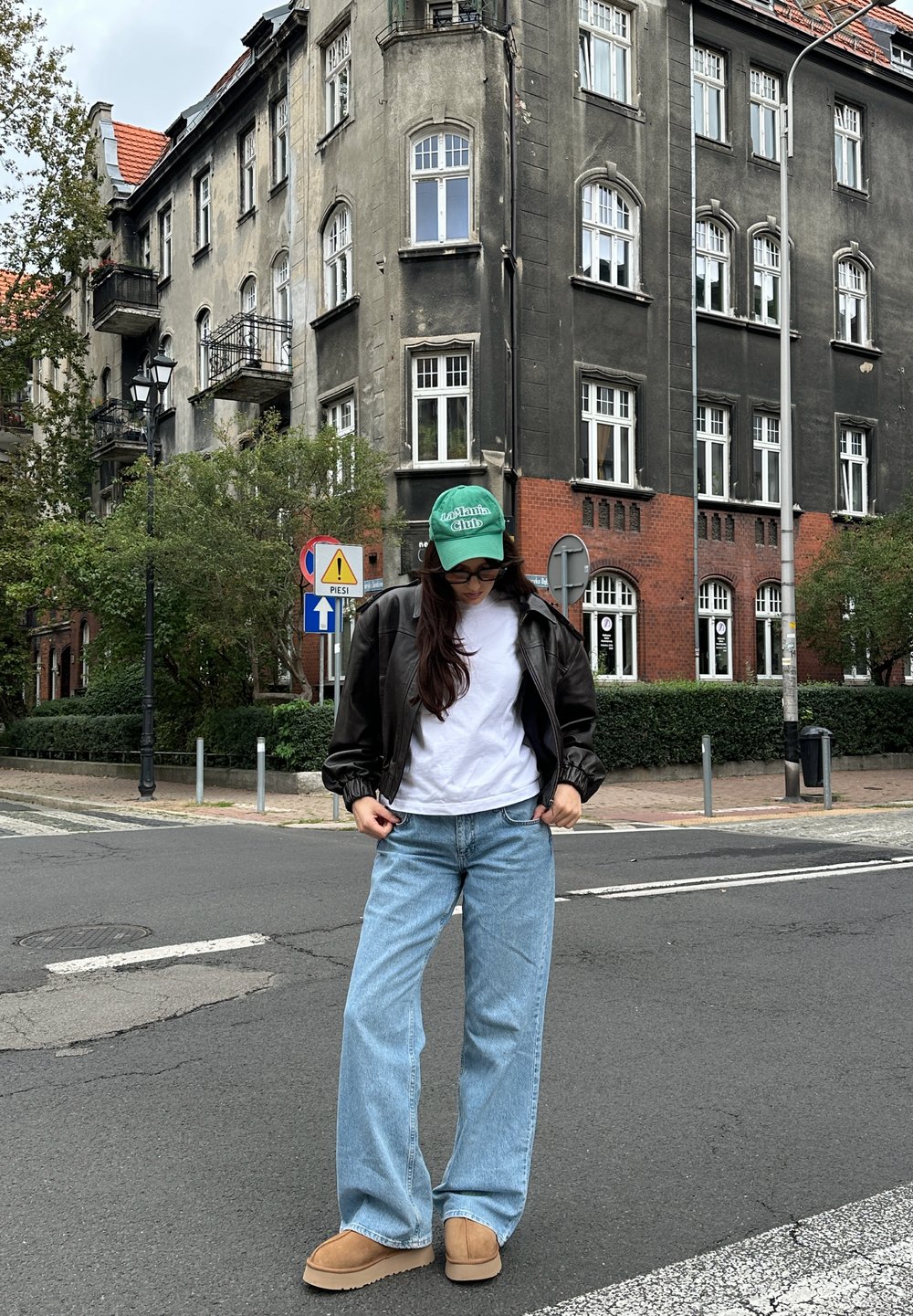 Model wearing a black leather bomber jacket, a white t-shirt, light blue wide-leg jeans, and tan slip-on shoes, complemented by a green cap, outdoors.