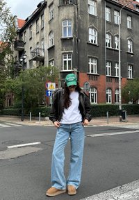 Model wearing a black leather bomber jacket, a white t-shirt, light blue wide-leg jeans, and tan slip-on shoes, complemented by a green cap, outdoors.