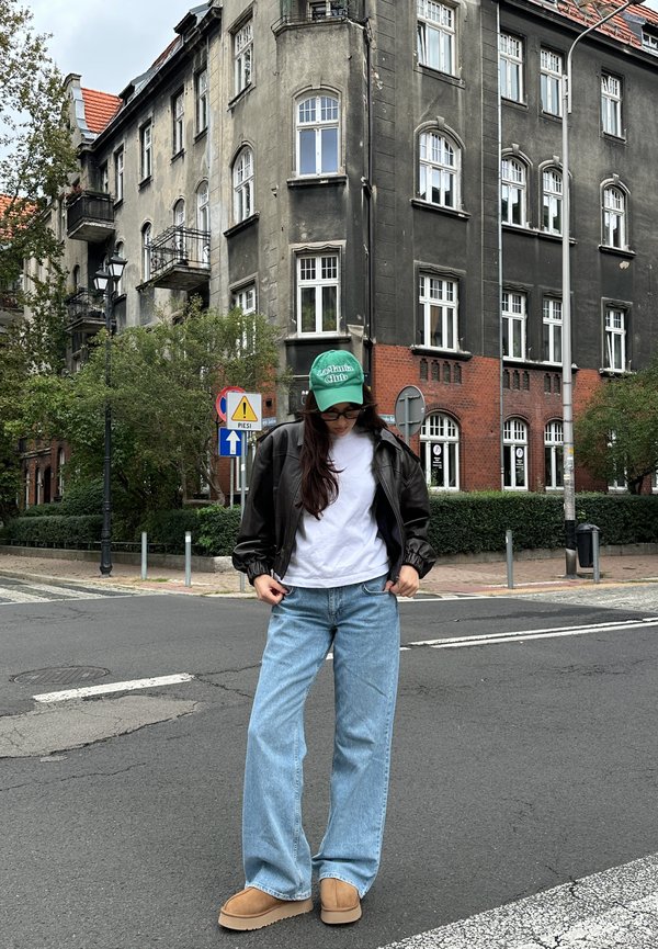Model fekete bőr bombázó dzsekiben, fehér pólóban, világoskék, széles szárú farmernadrágban és bézs slip-on cipőben, egy zöld sapkával a szabadban.
