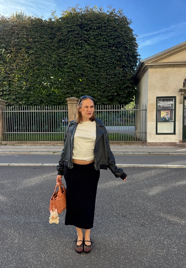 Fustă midi neagră, tricou alb scurt, jachetă din piele neagră și pantofi cu model. Ținând o geantă portocalie împletită cu un breloc pufos.