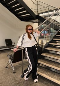 Tenue athlétique noir et blanc avec trois bandes, haut blanc court, pantalon noir et baskets. Présente un cadre intérieur minimaliste.