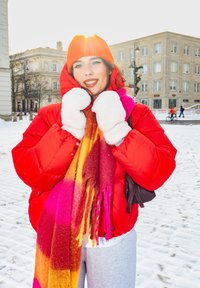 Νεαρή γυναίκα με φωτεινό κόκκινο μπουφάν, πορτοκαλί πλεκτό καπέλο, λευκά γάντια και πολύχρωμο κασκόλ στέκεται σε χιονισμένη αστική πλατεία την αυγή.