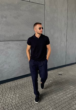 Camisa polo negra, pantalones sueltos azul marino y zapatillas oscuras. Gafas de sol con montura redonda. Paseando sobre un pavimento gris texturizado junto a una pared.