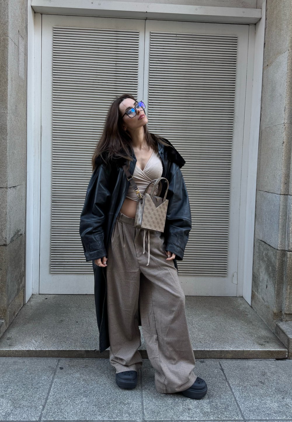 Femme portant un manteau en cuir noir surdimensionné, un haut beige enveloppant, un pantalon large à carreaux, des chaussures à plateforme noires et un sac bandoulière à motif, debout en plein air.