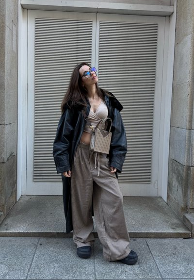 Mujer que lleva un abrigo de cuero negro de gran tamaño, blusa beige cruzada, pantalones anchos a cuadros, zapatos de plataforma negros y bolso cruzado con estampado, de pie al aire libre.