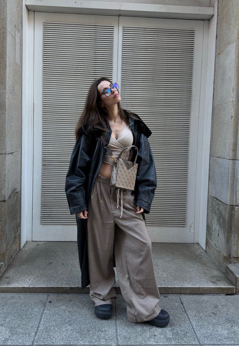 Γυναίκα που φοράει υπερμεγέθη μαύρο δερμάτινο παλτό, μπεζ τοπ τύπου wrap, πλατύριχα καρό παντελόνια, μαύρα παπούτσια με πλατφόρμα και τσάντα χιαστί με σχέδια, στέκεται σε υπαίθριο χώρο.