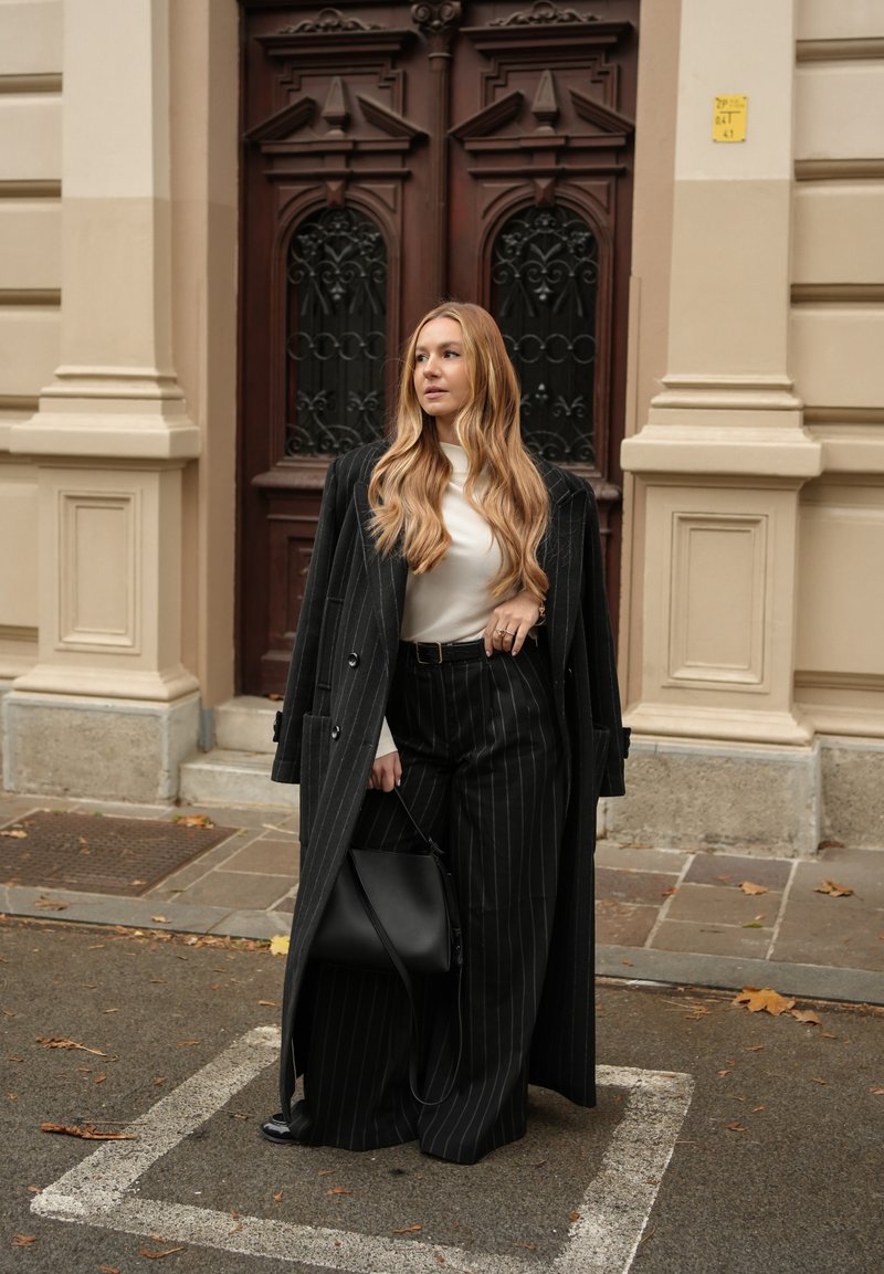 Frau mit langem blondem Haar trägt schwarzen Nadelstreifenmantel und Hose, weißes Oberteil, hält schwarze Handtasche, steht vor einer kunstvoll verzierten Holztür.