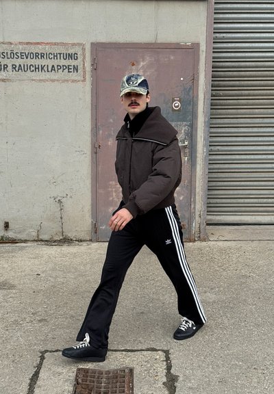 Chaqueta de plumas marrón con un cuello alto, pantalones de chándal negros de Adidas con franjas blancas y zapatillas negras. Fondo urbano con una puerta de metal.